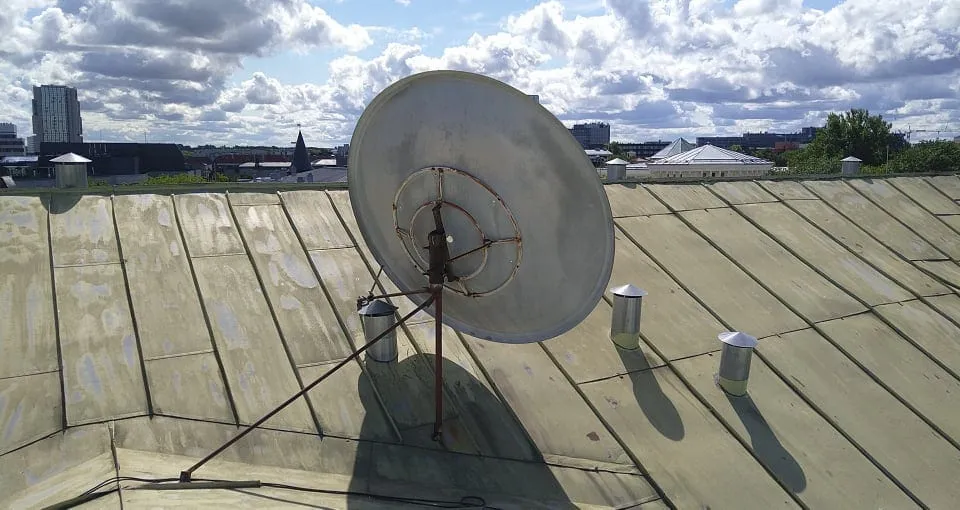 Suur satelliitantenn roostetanud mastil hoone plekk-katusel, taustal Tallinna linnavaade