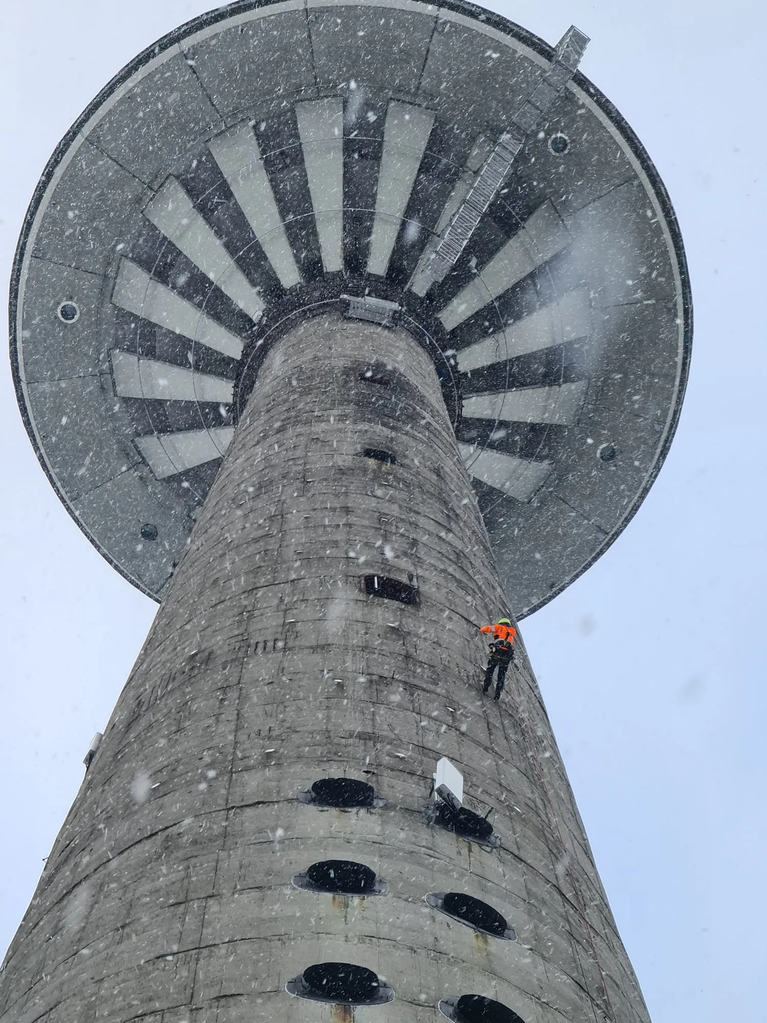 Köistehnik oranžis tööriietuses teletorni betoonsamba välisküljel lumesajus, kohal torni ketaskujuline vaateplatvorm
