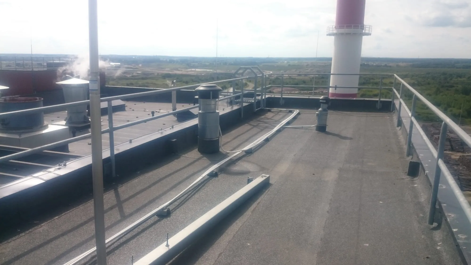 Tööstushoone katus piirete ja ventilatsioonitorudega, taustal punavalge korsten.